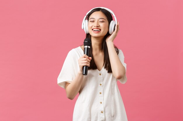 Beauty, people emotions and technology concept. Carefree happy asian girl using mobile phone karaoke application, singing in microphone, listen music in headphones, pink background.