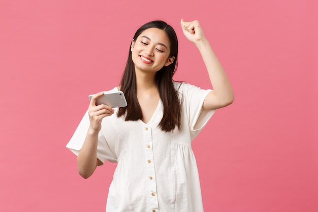 Beauty, people emotions and technology concept. Girl enjoying watching favorite k-pop music video on smartphone, dancing to beat, looking at mobile phone and chanting, pink background.