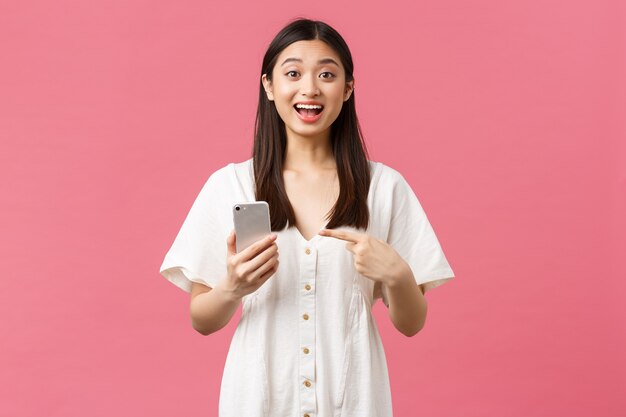美しさ、人々の感情と技術の概念。素晴らしいアプリケーションを見つけたようにスマートフォンで指を指している白いドレスを着た格好良いアジアの女の子を喜ばせ、携帯電話アプリをお勧めします