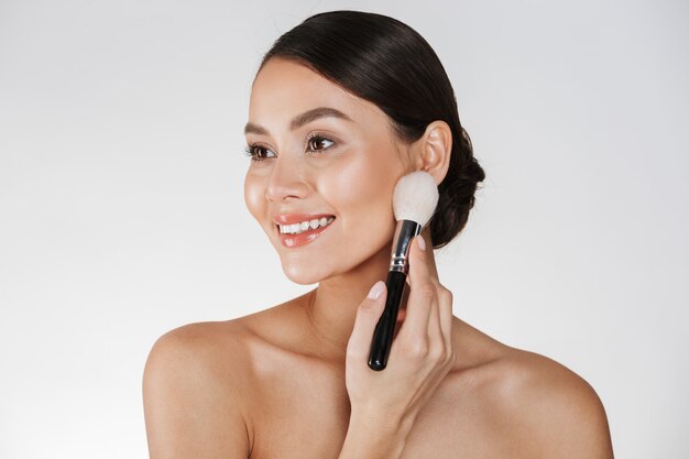 Beauty picture of attractive woman with perfect smile looking aside and applying makeup using brush, isolated over white