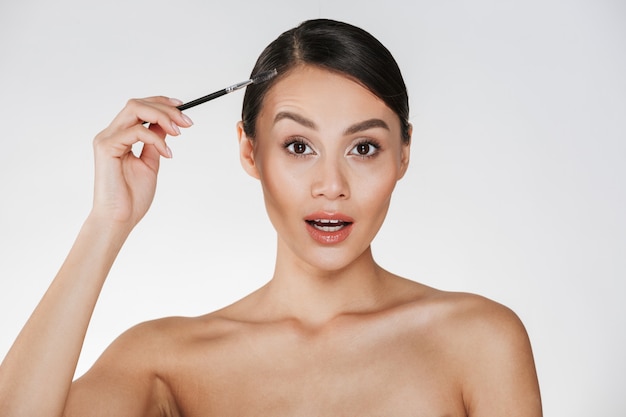 Beauty picture of gorgeous woman with hair in bun looking on camera and holding brush for eyebrows, isolated over white