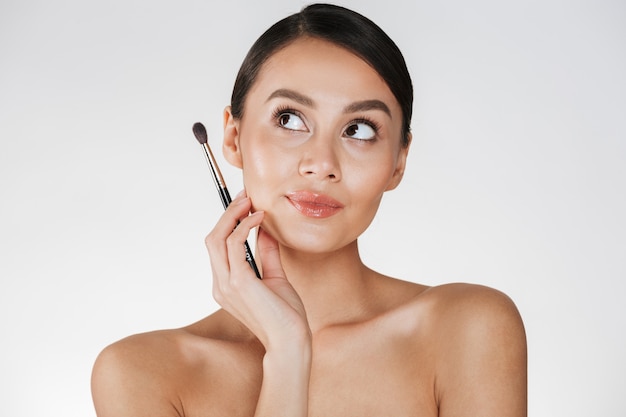 Beauty picture of gorgeous woman with hair in bun looking upward and holding brush for eyeshadow, isolated over white