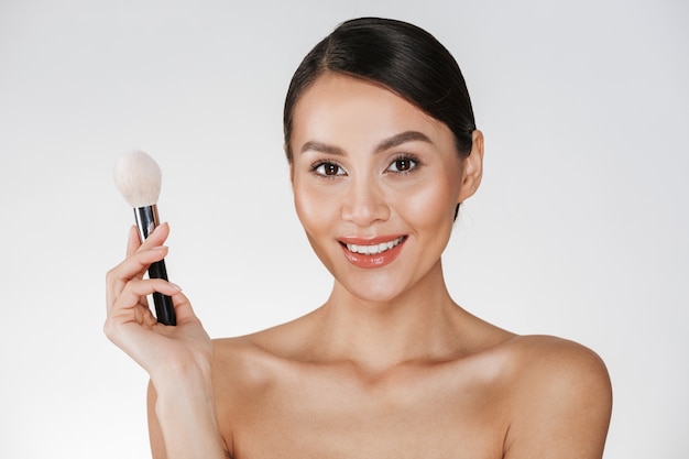 Beauty picture of pleased smiling girl with perfect skin looking on camera and holding makeup brush, isolated over white