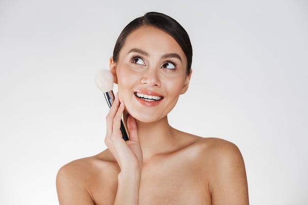 Beauty picture of pretty young woman with hair in bun looking upward and holding makeup brush close to her face, isolated over white