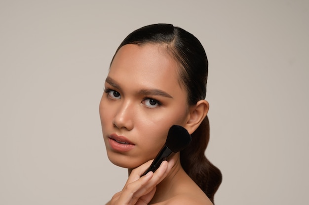 Beauty portrait of asian attractive sensual young woman pose holding makeup blusher brush on her cheek isolated over gray wall