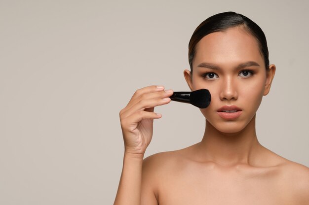 Beauty portrait of asian attractive sensual young woman pose holding makeup blusher brush on her cheek isolated over gray wall