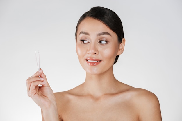 Beauty portrait of attractive candid dark-haired woman looking at small tweezers holding in her hand, isolated over white