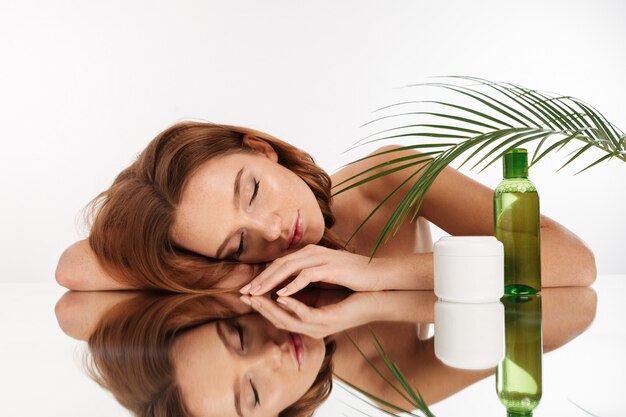 Beauty portrait of attractive ginger woman with long hair lying on mirror table with closed eyes near the bottle of lotion