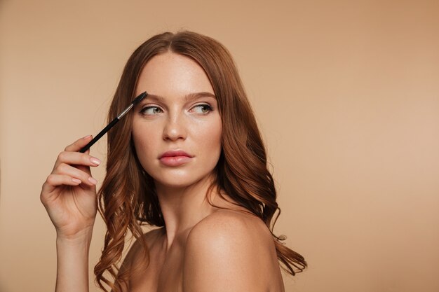 Beauty portrait of calm ginger woman with long hair looking away and posing sideways while combing her eyebrows with brush