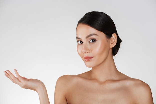 Beauty portrait of charming woman with brown hair in bun looking at camera and demonstrating something on her palm, isolated over white