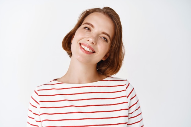 Beauty. Portrait of cheerful girl with short hairstyle, tilt head and smiling friendly, looking happy , standing against white wall