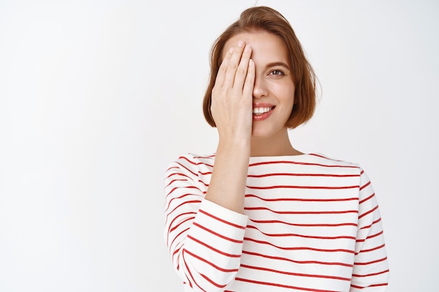 Beauty. Portrait of cute smiling woman cover half face with hand and looking happy, showing before after cosmetics skin care effect, standing against white wall