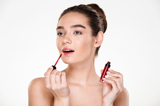 Beauty portrait of elegant half-naked lady with hair in bun applying red lipgloss looking aside