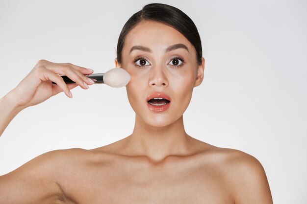 Beauty portrait of excited woman with perfect skin looking on camera and applying makeup using brush, isolated over white
