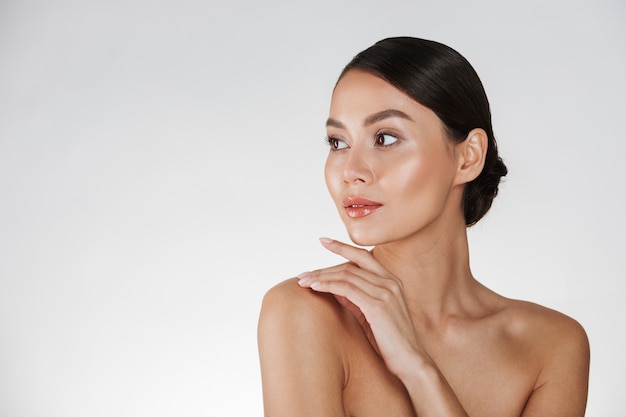 Beauty portrait of feminine young woman looking away and touching her nude shoulder, isolated over white