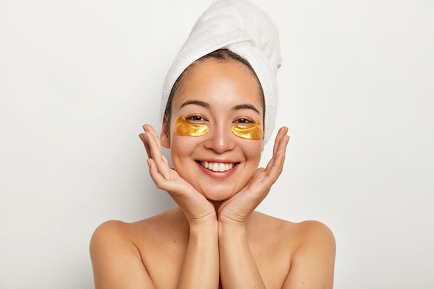 Beauty portrait of happy Asian girl keeps palms near face, looks positively, shows white perfect teeth, enjoys spa procedures, stands with wrapped towel on head