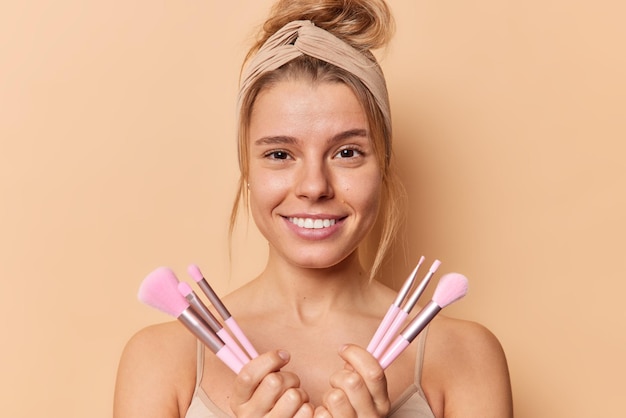 Free photo beauty portrait of lovely pleased young woman smiles happily holds cosmetic brushes for applying makeup wears headband and casual t shirt looks directly at camera isolated over brown background