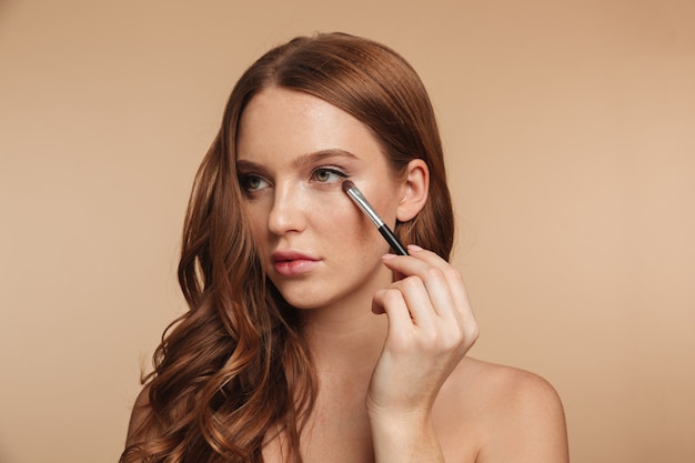 Free photo beauty portrait of mystery smiling ginger woman with long hair looking away while applying cosmetics with brush for eyeshadow