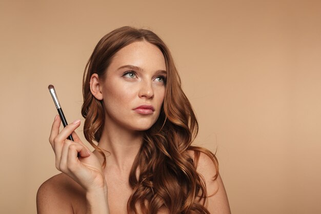 Beauty portrait of pensive ginger woman with long hair looking up while holding cosmetics brush