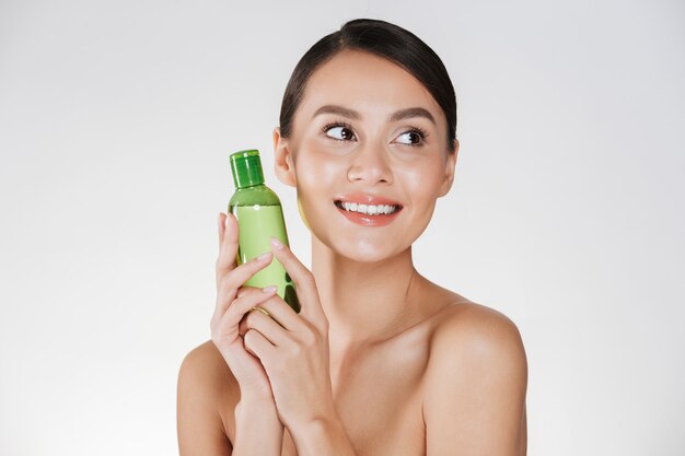 Beauty portrait of pleased dark-haired woman with clean healthy skin holding lotion for removing makeup and looking aside, isolated over white