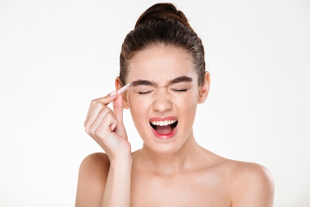 Beauty portrait of sensual brunette woman with hair in bun screaming in pain while plucking eyebrows with tweezers