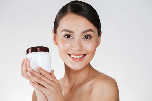 Beauty portrait of smiling brunette woman with soft healthy skin holding bank with face cream, and looking on camera isolated over white