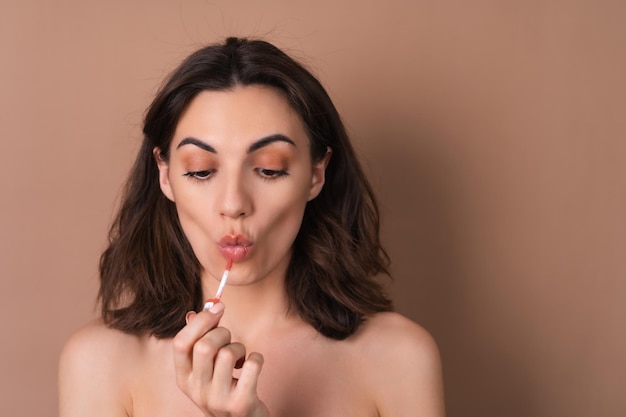 Beauty portrait of topless woman with perfect skin and natural makeup on beige background holding brown chocolate gloss lipstick