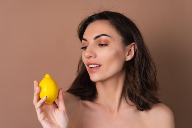 Beauty portrait of topless woman with perfect skin and natural makeup on a beige background holds citrus lemon vitamins c for skin