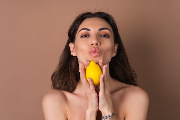 完璧な肌とベージュの背景に自然なメイクでトップレスの女性の美しさの肖像画は、肌のための柑橘類のレモンビタミンCを保持しています