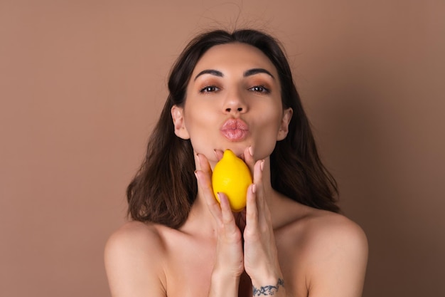 Beauty portrait of topless woman with perfect skin and natural makeup on a beige background holds citrus lemon vitamins c for skin