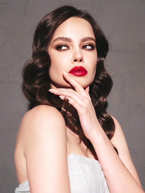 Beauty portrait of young brunette woman with evening makeup and perfect clean skin Sexy model with curly hair posing in studio near dark wall With red bright natural lips In silver dress