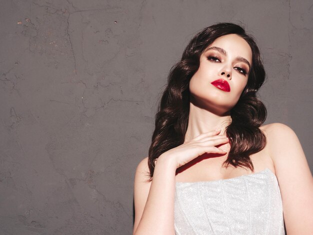 Beauty portrait of young brunette woman with evening makeup and perfect clean skin Sexy model with curly hair posing in studio near dark wall With red bright natural lips In silver dress