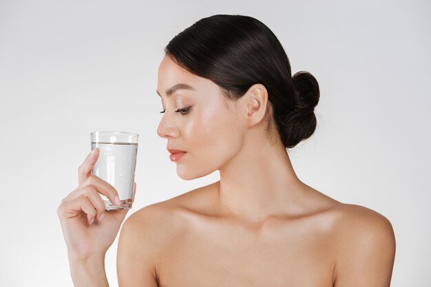 Beauty portrait of young happy woman with hair in bun looking at transparent glass of still water holding in hand, isolated over white