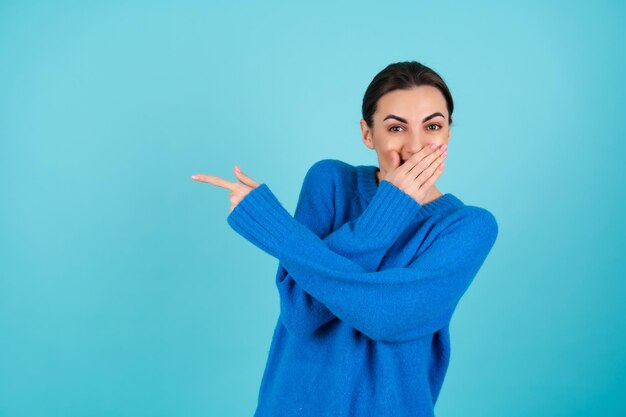 Beauty portrait of a young woman in a blue knitted sweater and natural day makeup, smiling cheerfully and pointing her finger to the left to an empty space