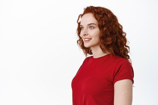 Beauty. Portrait of young woman with red curly hair and pale smooth skin looking left at empty copy space smiling happy, white wall