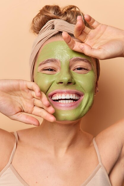 미용 절차 및 피부 관리 개념입니다. 쾌활한 여성은 얼굴에 손을 대고 미소를 지으며 긍정적으로 하얀 치아가 얼굴 치료를 즐긴다는 것을 보여줍니다. 녹색 영양 마스크는 실내에서 머리띠를 하고 포즈를 취합니다