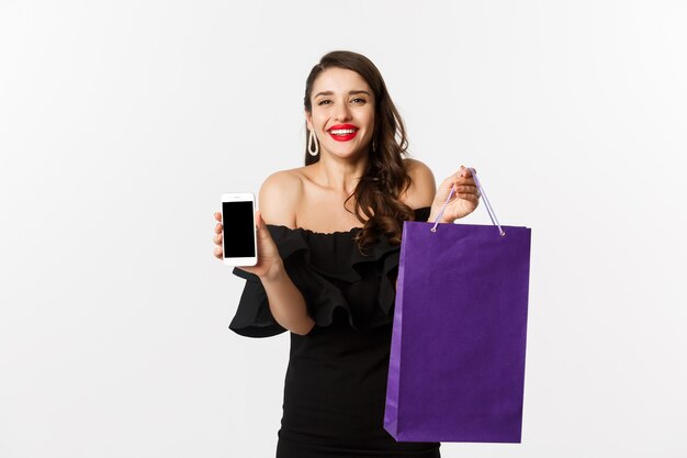 Beauty and shopping concept. Beautiful and stylish woman showing mobile phone screen and bag, buying online, standing over white background