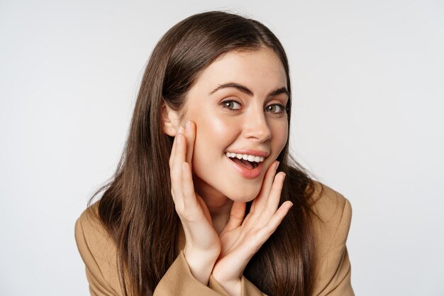 Beauty and skin care. Close up portrait of beautiful woman looking in mirror and touching her clear glowing face, standing over white background.