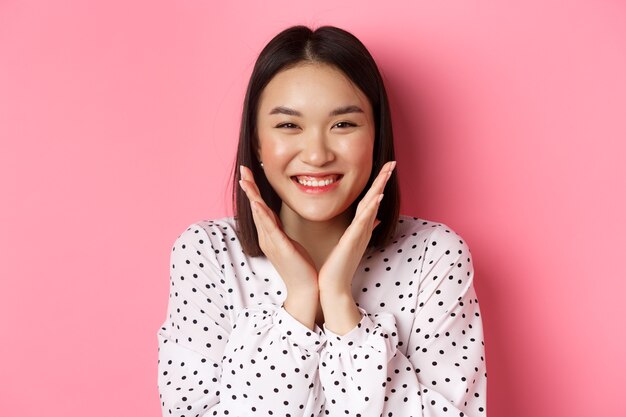 아름다움과 피부 관리 개념입니다. 깨끗하고 완벽한 얼굴을 보여주는 귀여운 아시아 여성의 클로즈업, 웃고, 카메라를 보고 행복해하며, 분홍색 배경 위에 서 있습니다