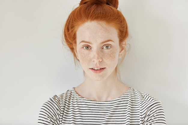 Free photo beauty and skin care concept. close up isolated shot of cute stylish young redhead woman with freckles and hair bun looking with charming smile, standing at white wall, wearing sailor shirt