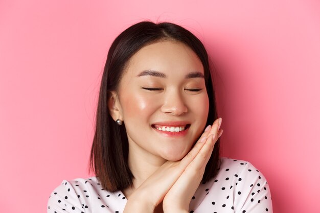 Beauty and skin care concept. Headshot of adorable and dreamy asian woman close eyes, smiling nostalgic, standing against pink background