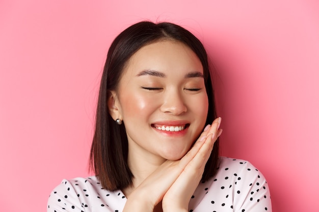 Beauty and skin care concept. Headshot of adorable and dreamy asian woman close eyes, smiling nostalgic, standing against pink background