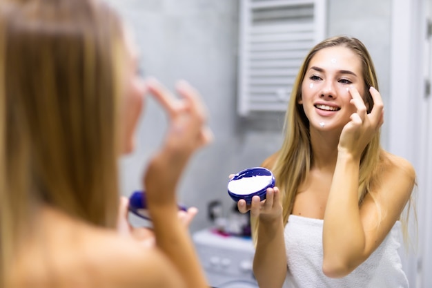 Beauty, skin care and people concept - close up of young woman applying cream to face and looking to mirror at home bathroom