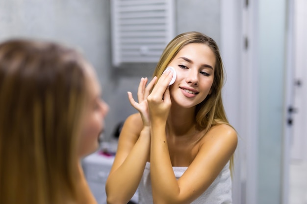 아름다움, 피부 관리 및 사람 개념 - 화장실에서 얼굴을 씻기 위해 면 디스크에 로션을 바르는 웃고 있는 젊은 여성