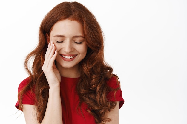 Beauty and skin care. Redhead woman lovely smiling, looking down flirty and touching gentle natural facial skin, standing in t-shirt against white background.