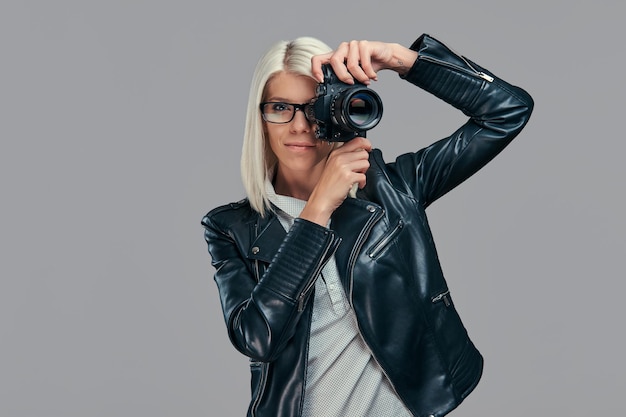 Beauty smart blonde female photographer in a white blouse and black leather jacket taking a photo at a studio. Isolated on a gray background.