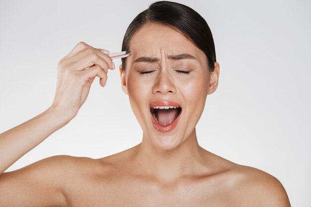 Beauty of stressed young woman with brown hair screaming in pain while plucking eyebrows using tweezers, isolated over white