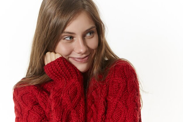 Beauty, style and fashion concept. Charming playful young female in cozy long sleeved sweater holding hand on her cheek and looking away with curious smile, listening attentively to interesting story