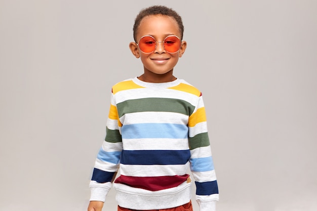 Beauty, style and fashion concept. Picture of cheerful fashionable African boy posing isolated wearing stylish striped sweater and trendy round pink sunglasses, smiling happily