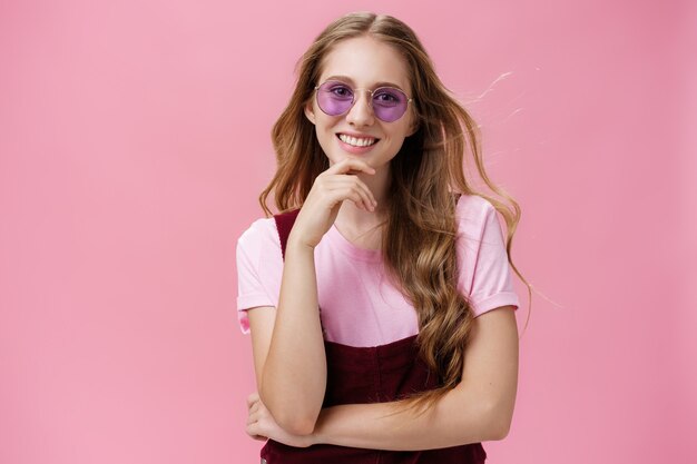 Beauty, style and fashion concept. Portrait of charming confident european female with natural wavy hair in sunglasses and overalls touching chin amused and smiling broadly against pink wall.